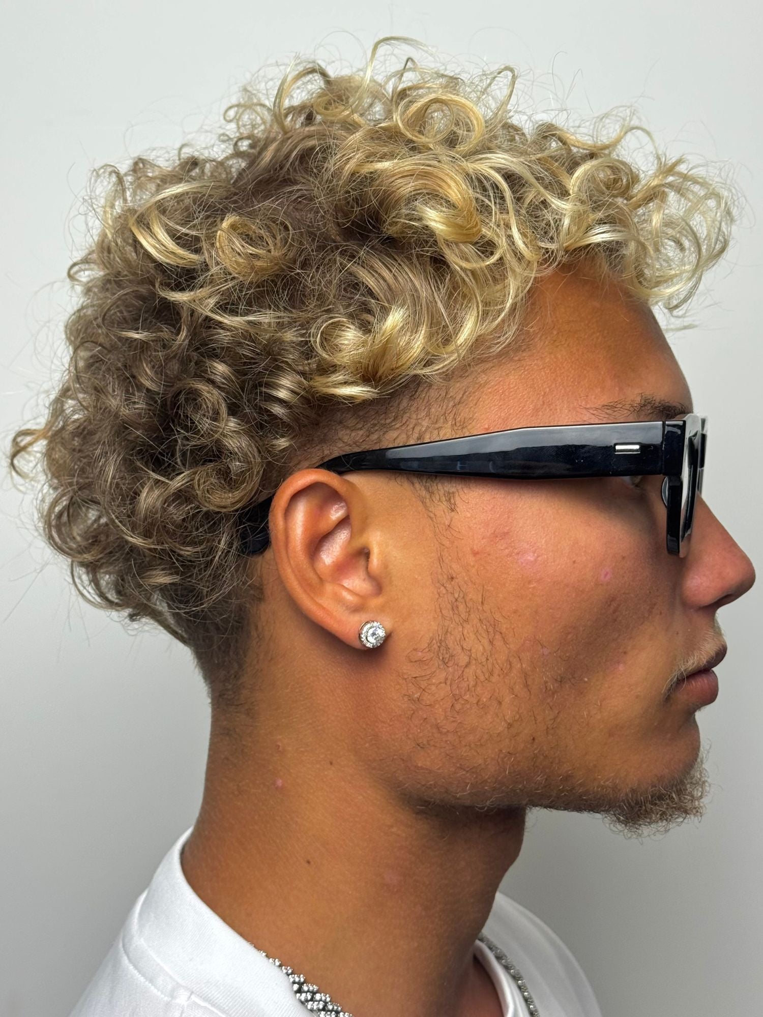 Man with curly blonde hair wearing sunglasses and stud earrings, showcasing a stylish profile view.
