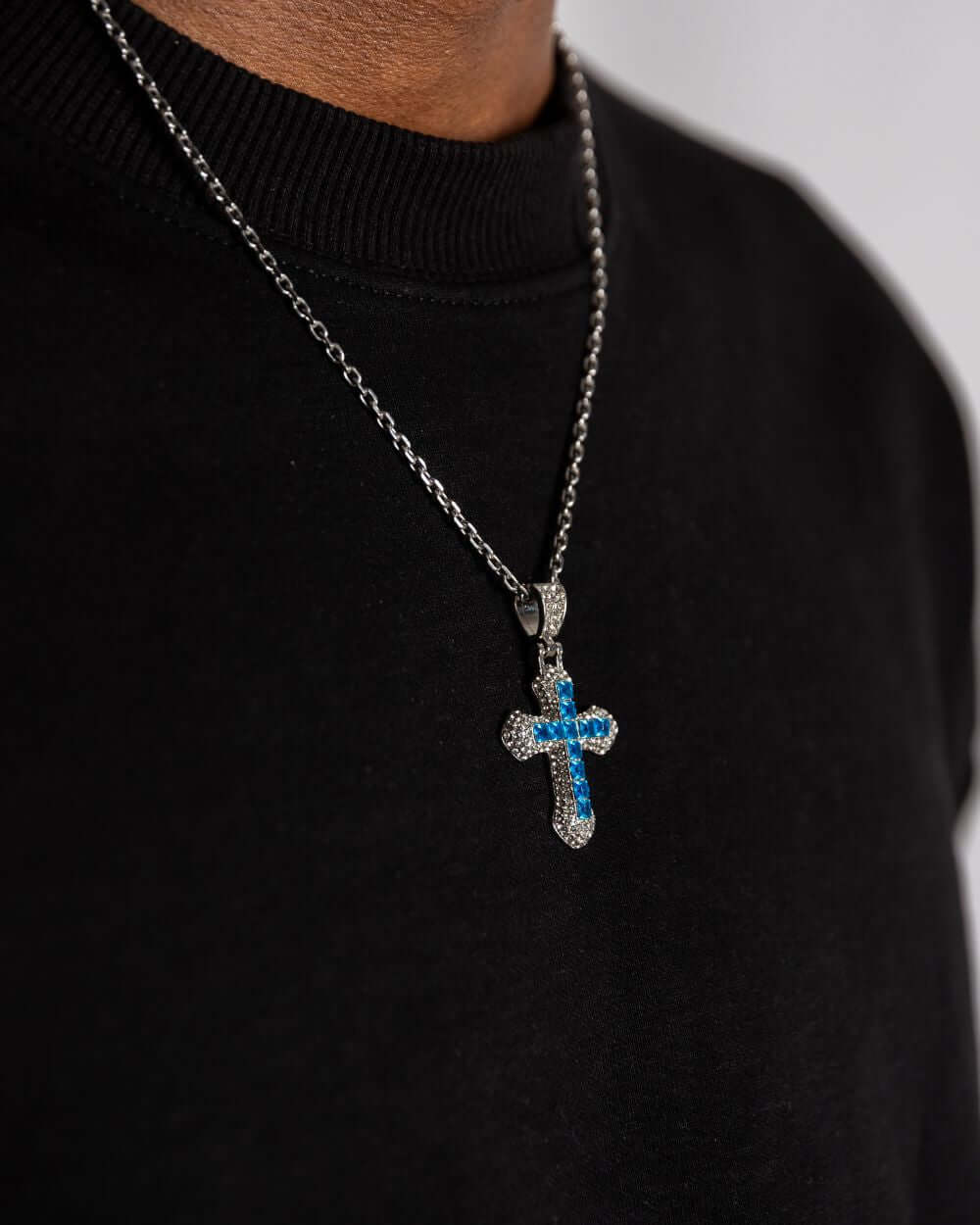 Blaues Kreuz-Anhänger aus Silber, ICED OUT, mit blauen Steinen, an einer Kette auf einem schwarzen Shirt getragen.