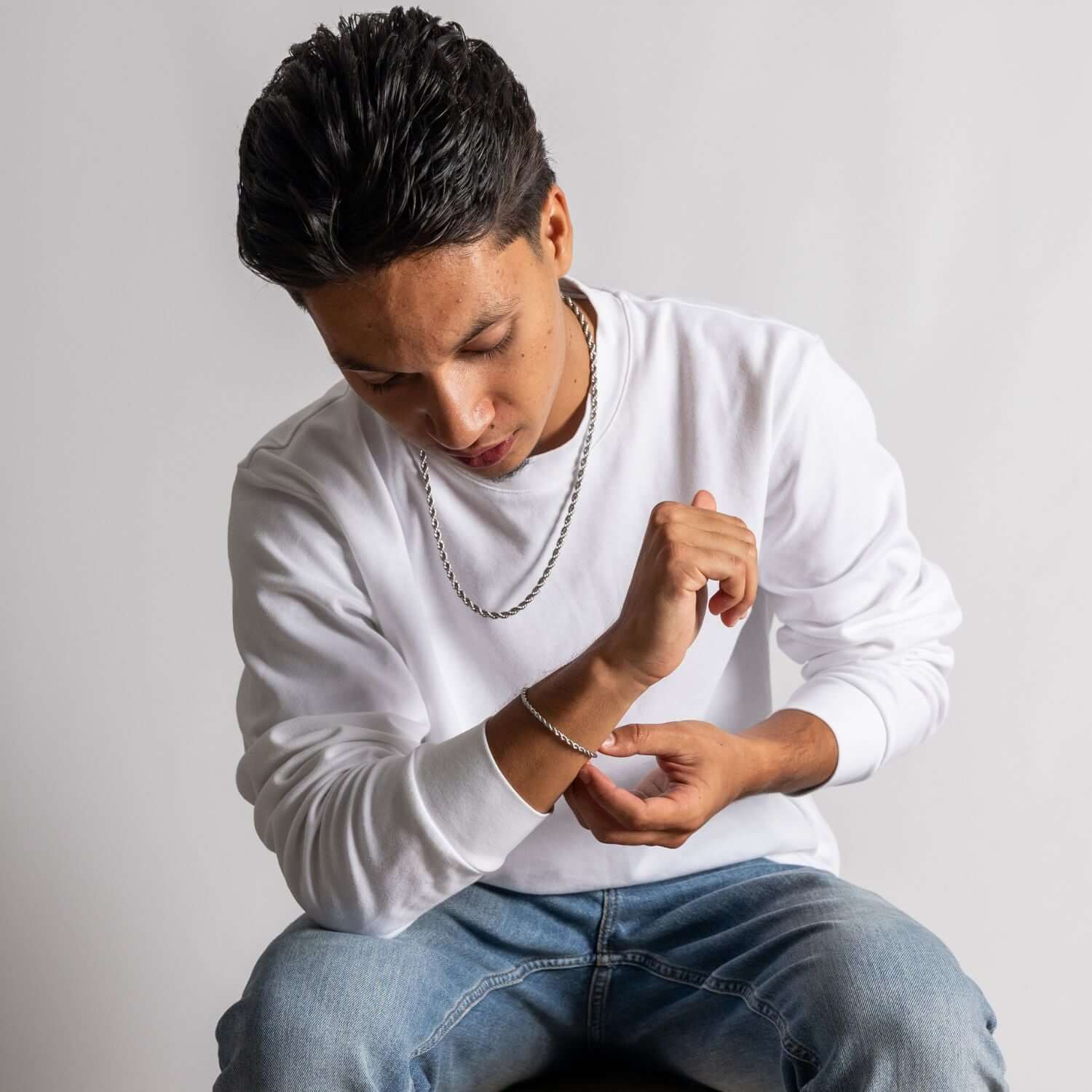Mannelijk model in witte sweater en jeans met zilveren ketting en armband, studioportret met neutrale achtergrond