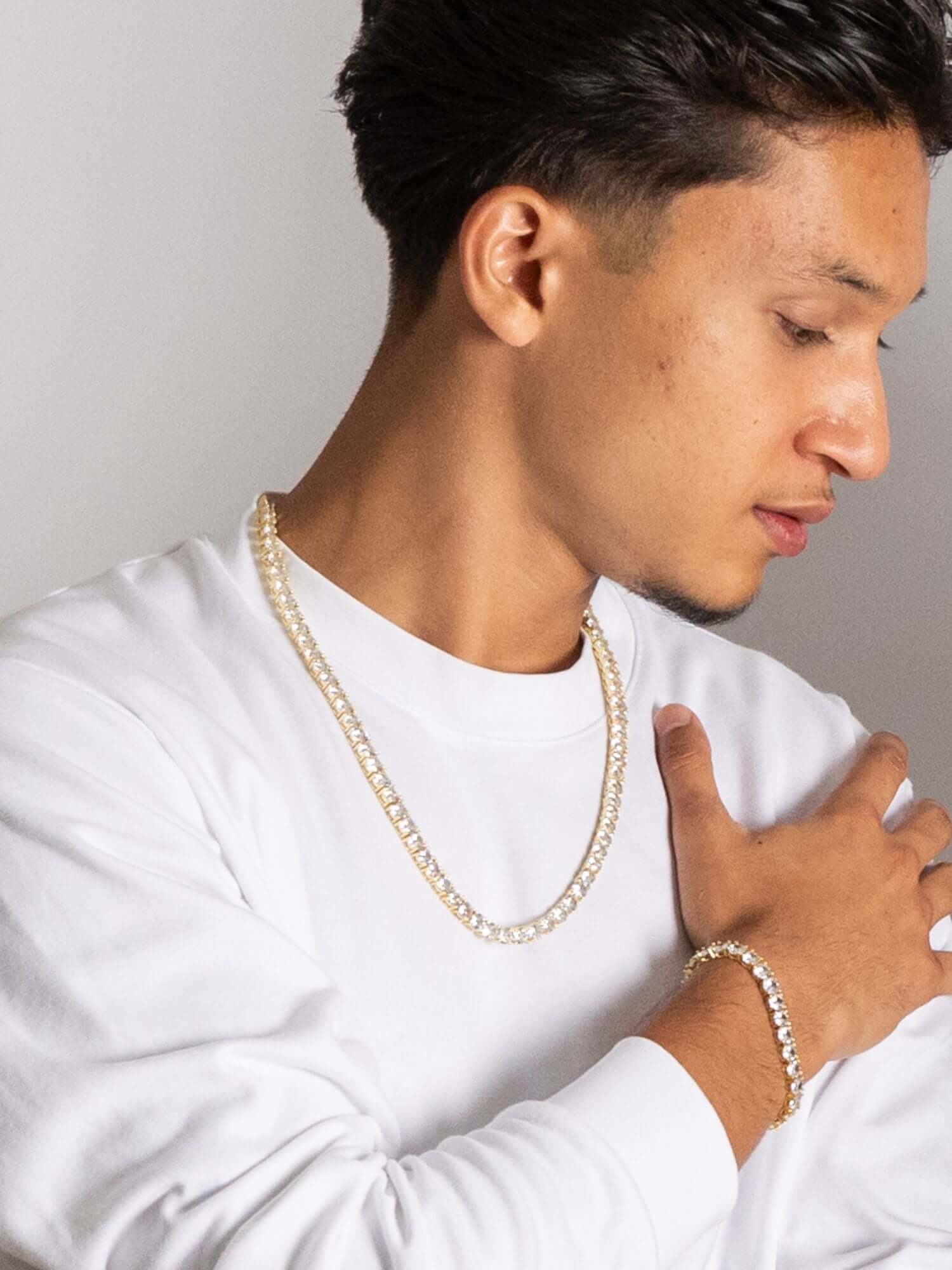Gouden tennis ketting en armband worden gedragen door een man in casual witte outfit.