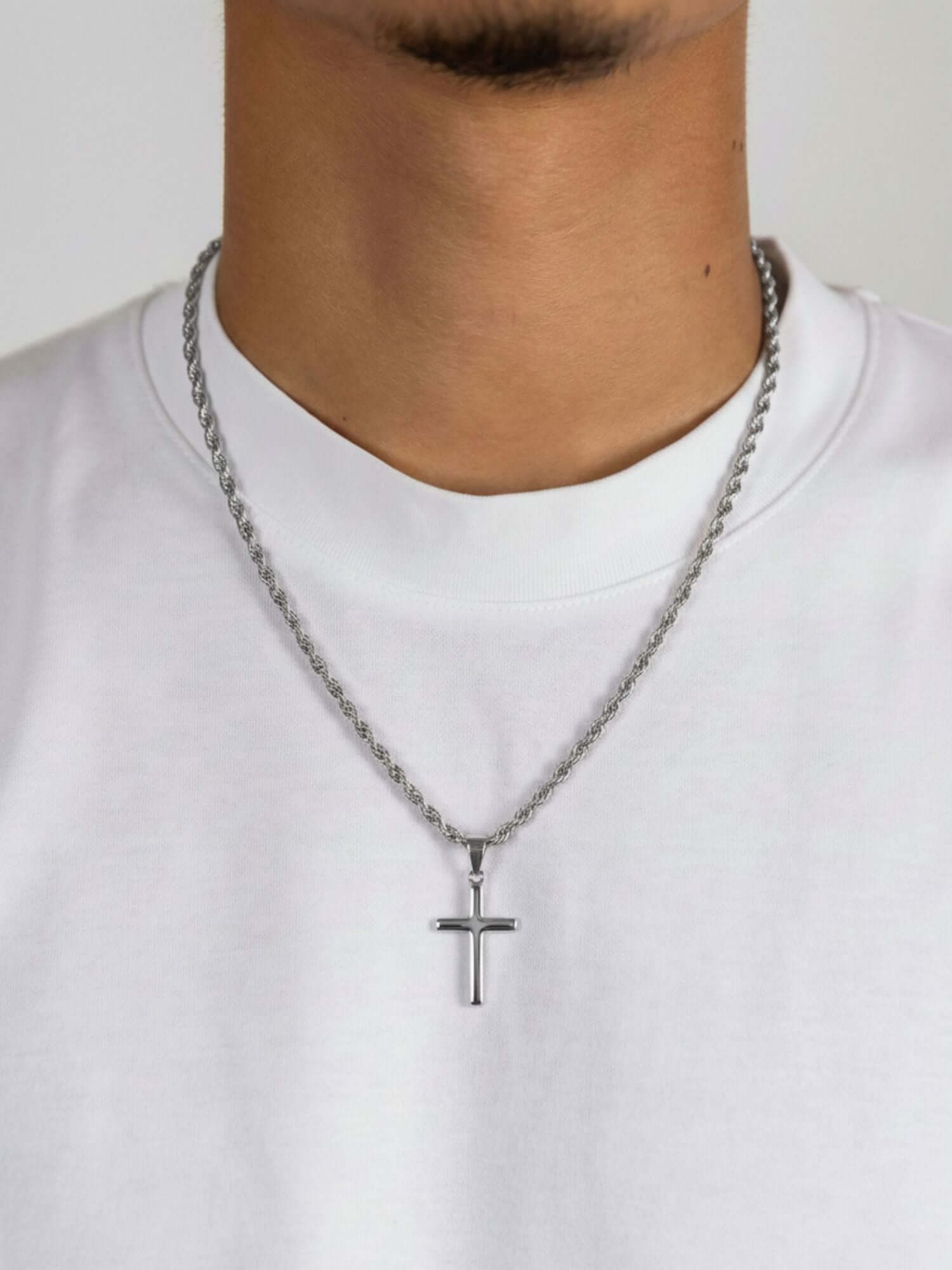 Silverfärgat korshänge buren av en man, kombinerat med ett silverfärgat halsband på en vit t-shirt.
