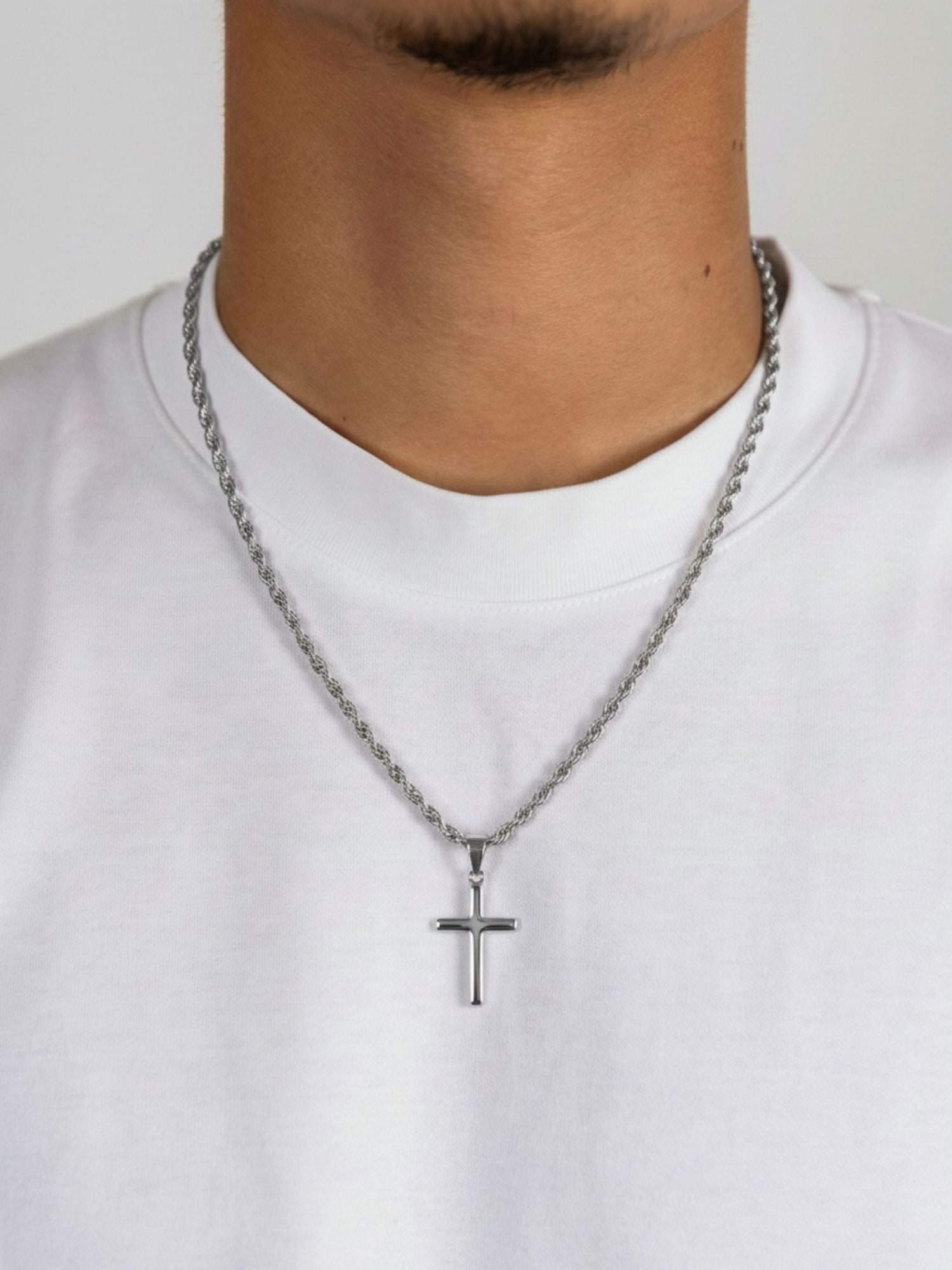 Zilveren kruis hanger gedragen door man, gecombineerd met een zilverkleurige ketting op een witte t-shirt.
